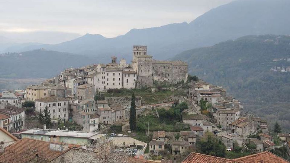 Veduta panoramica di un borgo collinare con case in pietra e tetti in coppi, disposto a terrazze attorno a un castello medievale con torre centrale. Sullo sfondo si vedono rilievi montuosi e valli boscose sotto un cielo nuvoloso.