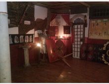 Interno di una sala espositiva a tema romano, con pareti in mattoni e pannelli rossi decorati, scudi e armature in mostra e un piccolo altare o piedistallo su pavimento in legno. Sul fondo si vedono drappi rossi, una grata a finestra e teche o pannelli con oggetti e illustrazioni.