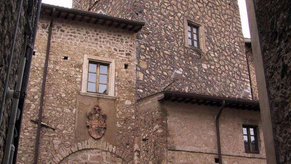 Torre medievale in pietra con finestre ad arco nella parte alta, vista dal basso in un vicolo stretto tra edifici in muratura. Alla base si vede un grande portale ad arco con stemma sopra e piccole finestre incorniciate.