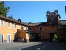 Cortile lastricato circondato da edifici intonacati color ocra con finestre e tetti in coppi, con un grande portale ad arco che conduce all’esterno. Sullo sfondo si vede una struttura tipo campanile con due piccole campane e un orologio sotto un cielo sereno.
