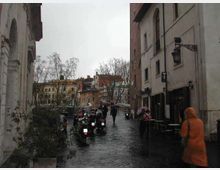 Strada urbana in pavé bagnato in un centro storico, fiancheggiata da edifici alti con facciate chiare e lampioni a parete. Sul lato destro si vedono tavolini all’aperto e una fila di scooter parcheggiati, con alberi spogli e palazzi sullo sfondo.