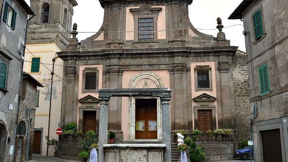 Facciata di una chiesa storica in pietra e intonaco rosa, con portale centrale incorniciato, due ingressi laterali e campanile sul lato sinistro, affacciata su una piccola piazza. In primo piano si vede una fontana ottagonale in pietra al centro della pavimentazione circolare, circondata da edifici residenziali.