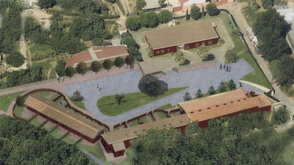 Veduta aerea di un complesso rurale con più edifici rettangolari dai tetti rossi disposti attorno a una grande area pavimentata. Al centro ci sono aiuole verdi con alberi e vialetti curvi, circondati da campi e vegetazione.