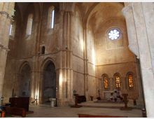 Interno di una chiesa in pietra con alte arcate a sesto acuto, pilastri massicci e volte a crociera. Sul fondo si vede l’area dell’altare sopraelevata, illuminata da finestre strette e da un rosone circolare sulla parete.
