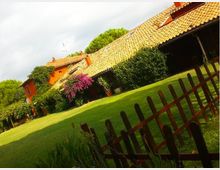 Casa di campagna con tetto in tegole e facciata arancione, circondata da un ampio prato verde e cespugli fioriti. In primo piano si vede una staccionata in legno lungo il bordo del giardino.