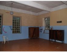 Interno di una stanza spoglia con pavimento in piastrelle marroni, pareti bicolori beige e azzurre e soffitto alto con lampada a sospensione. Due finestre con inferriate e alcuni mobili in legno lungo le pareti, con graffiti visibili sulle superfici.