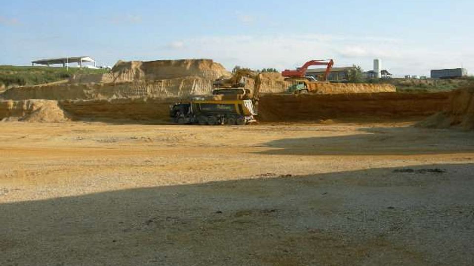 Grande cava a cielo aperto con pareti di terra stratificata e ampie superfici di sabbia e ghiaia; sullo sfondo si vedono macchinari da scavo e depositi di materiale sotto un cielo aperto.