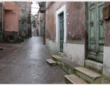 Vicolo stretto con pavimentazione in pietra, fiancheggiato da edifici antichi con intonaco usurato e portoni in legno verde con piccoli gradini d’ingresso. In alto si vedono balconi in ferro e cavi sospesi lungo la strada che curva tra le case.