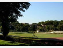 Campo sportivo all’aperto con ampia area erbosa centrale e pista ovale rossa attorno, con una piccola tribuna metallica a bordo campo. Sullo sfondo si vedono alberi alti e colline con edifici in muratura, sotto un cielo sereno.