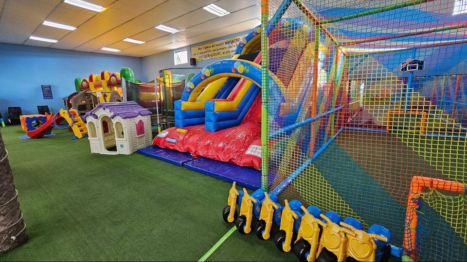 Area giochi interna per bambini con pavimento in erba sintetica, gonfiabili colorati con scivolo e un grande percorso gioco a rete con trampolini. Sullo sfondo si vedono casette e piccoli scivoli, sotto un soffitto con pannelli e luci al neon.