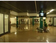 Ampia hall interna con pavimento lucido in piastrelle, soffitto a listelli scuri e pilastri verdi, con panche metalliche e alcune piante in vaso. Sulle pareti sono presenti poster e, in alto, un monitor di sorveglianza; sul fondo si vedono porte e corridoi di accesso.