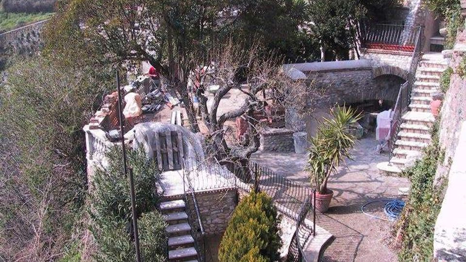 Cortile terrazzato in pietra su un pendio, con scale, parapetti in ferro e un vialetto curvo tra alberi e cespugli. Sullo sfondo si vedono case in pietra addossate l’una all’altra, tipiche di un borgo storico.