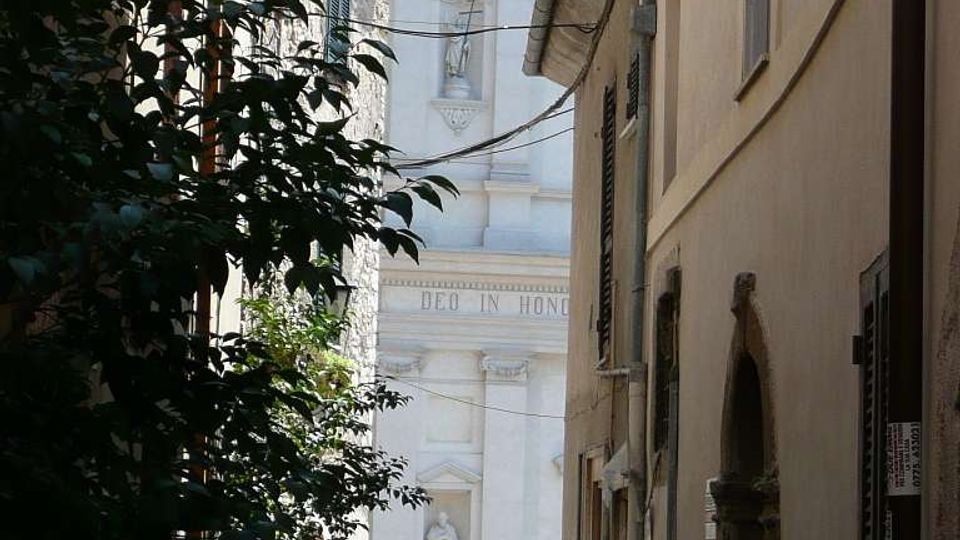 Vicolo stretto in pietra tra edifici alti con facciate intonacate e persiane, con alberi e cavi sospesi lungo la strada. In fondo si vede la facciata bianca di una chiesa in stile classico, con timpano, nicchie e statue.