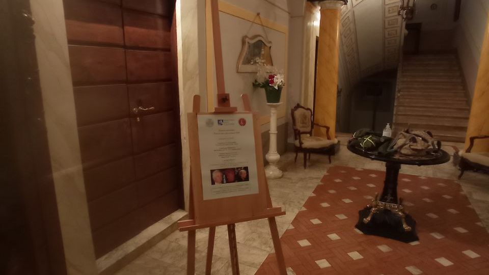 Ingresso interno di un edificio storico con pavimento in marmo e tappeto di piastrelle rosse a rombi, porta in legno imbottita e cavalletto con cartello informativo. Sul fondo un arco affrescato con stemma conduce a una scalinata, con tavolino rotondo, sedie e lampadario.