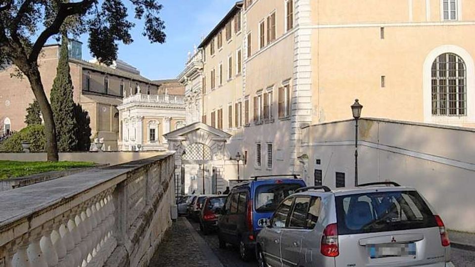 Strada in sampietrini in discesa fiancheggiata da un alto edificio storico intonacato color ocra con finestre regolari e da un muro in pietra con alberi sopra. Auto parcheggiate lungo il lato destro e, in fondo, un portale ad arco che conduce verso un complesso architettonico monumentale.