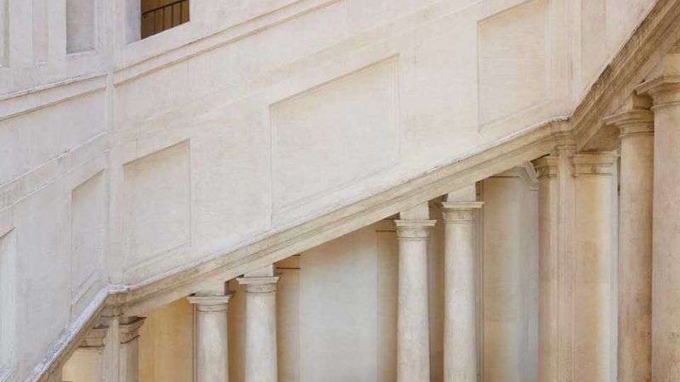 Interno di un edificio storico con ampia scalinata in pietra chiara che sale in diagonale, sostenuta da una fila di colonne. Pareti intonacate con riquadri decorativi e aperture superiori con ringhiere affacciano sul vano scala.
