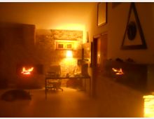 Ampio soggiorno con pareti in pietra e due camini accesi, con illuminazione calda e soffusa. Al centro c’è un tavolo con sedie e oggetti, e sul fondo si vede una piccola finestra incassata nel muro.