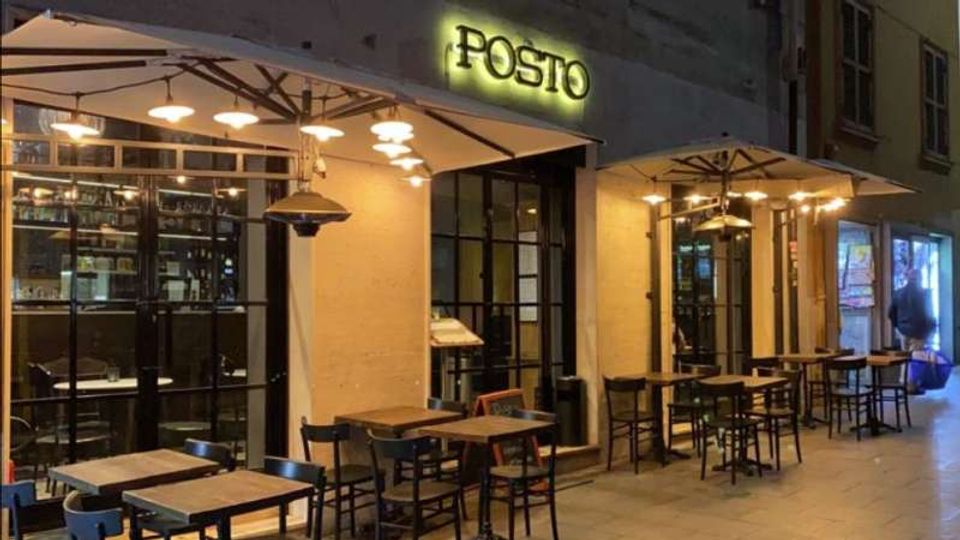 Facciata di un ristorante con insegna luminosa “POSTO”, grandi vetrate e veranda esterna con tavolini e sedie. Ampi ombrelloni con lampade pendenti illuminano il marciapiede in una strada urbana.