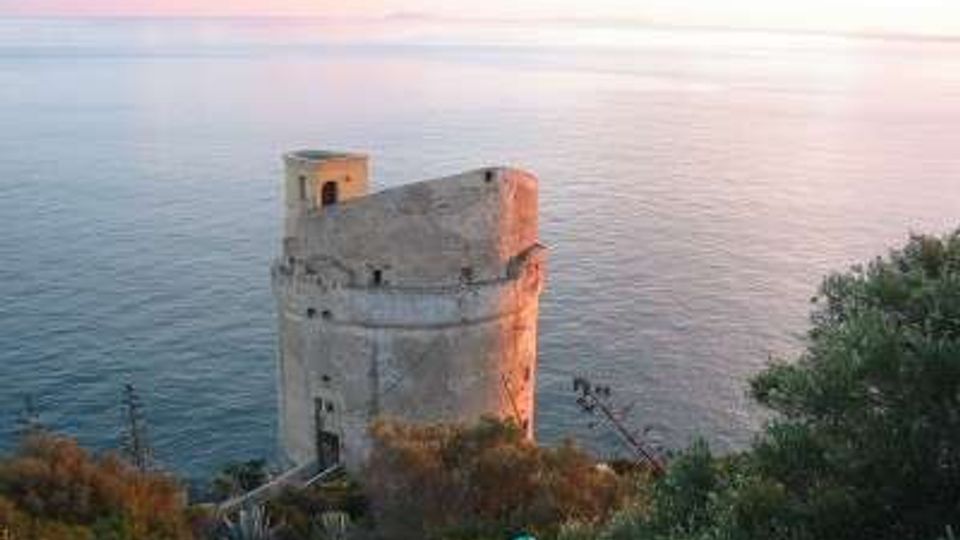 Torre costiera in pietra su un promontorio roccioso, circondata da vegetazione mediterranea e affacciata sul mare calmo. Sullo sfondo si vede l’orizzonte marino con cielo al tramonto.
