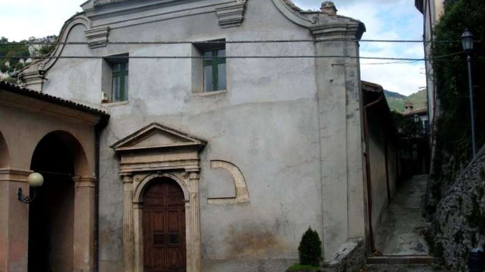 Facciata di una piccola chiesa in intonaco chiaro con timpano curvilineo, croce in sommità e portale in pietra con porta in legno. Davanti c’è un cortile lastricato e, sul lato destro, una stretta salita tra edifici che conduce verso l’alto.