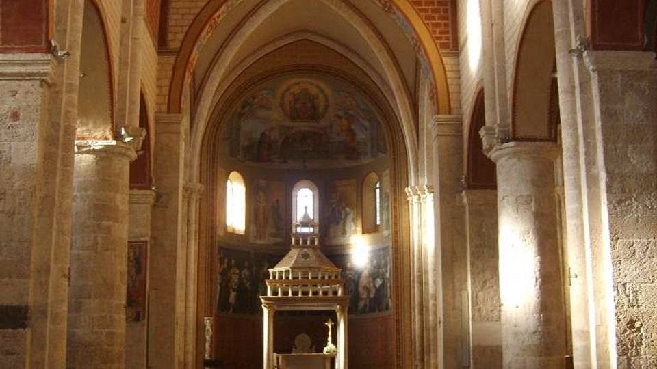 Interno di una chiesa in stile romanico-gotico con navata centrale, alte arcate in pietra e soffitto a capriate in legno. In fondo si vede l’altare sotto una struttura a baldacchino, affreschi sull’abside e file di sedie disposte lungo il pavimento decorato a motivi geometrici.