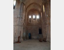 Interno di una chiesa in pietra con navata alta e volte a crociera, grandi pilastri e pareti spoglie. Sul fondo si vedono strette finestre ad arco che lasciano entrare luce, una porta e una scalinata laterale con poche panche.