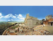 Veduta panoramica di un castello medievale in pietra con mura merlate e torri, affacciato su una terrazza con parapetti e vasi. In primo piano si vedono tetti in coppi e, sullo sfondo, una valle collinare verde sotto un cielo azzurro con nuvole.