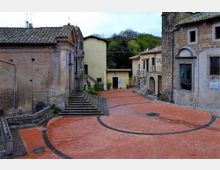 Piccola piazza lastricata in mattoni rossi con linee curve scure, circondata da edifici in pietra e intonaco e una scalinata laterale; sullo sfondo si vedono case basse e alberi su una collina.