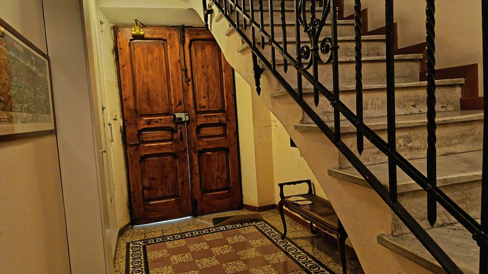 Ingresso interno di un edificio con grande portone in legno e pavimento a scacchiera con bordo decorato. A destra una scala in marmo con corrimano in ferro battuto e una piccola panca sotto le scale.