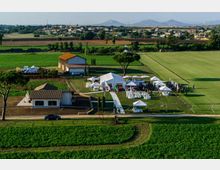 Veduta aerea di una tenuta in campagna con edifici bassi e un prato recintato allestito con grandi tensostrutture bianche, gazebo e file di sedie. Intorno si estendono campi coltivati e, sullo sfondo, un piccolo centro abitato e montagne lontane.