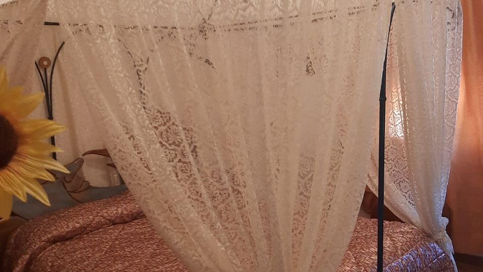 Camera da letto con letto matrimoniale coperto da un baldacchino in pizzo bianco fissato a una struttura metallica. Pareti chiare, pavimento in legno e finestra con tende arancioni sullo sfondo.