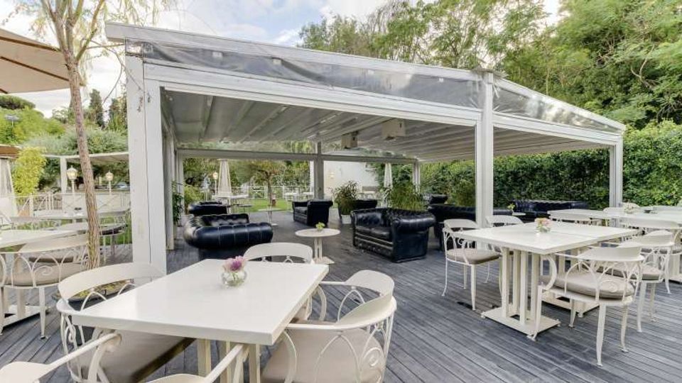 Terrazza all’aperto con pergolato bianco e copertura trasparente, pavimentazione in legno e tavoli bianchi con sedie coordinate. Sul fondo sono presenti divani scuri da lounge e siepi e alberi che circondano l’area.