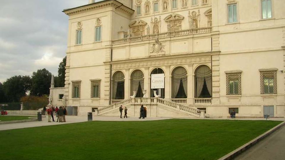 Facciata di un grande palazzo storico in stile classico, con decorazioni in rilievo, finestre alte e una loggia ad archi al piano terra. Davanti all’ingresso centrale con scalinata e balaustra si estende un ampio prato verde, con vialetti e alberi sullo sfondo.