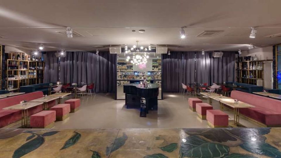 Interno di un lounge bar moderno con bancone centrale in pietra scura, lampadario a sfere luminose e scaffalature con bottiglie lungo le pareti. Ai lati sono presenti tavoli rettangolari con sedute imbottite rosa e tende scure che separano l’area del bar.