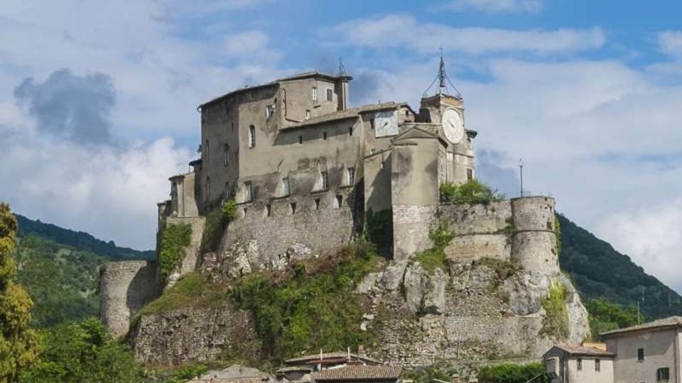 Castello medievale in pietra arroccato su uno sperone roccioso, con mura fortificate, torrioni e una torre con orologio. In primo piano si vedono i tetti di un borgo, con colline verdi e cielo parzialmente nuvoloso sullo sfondo.