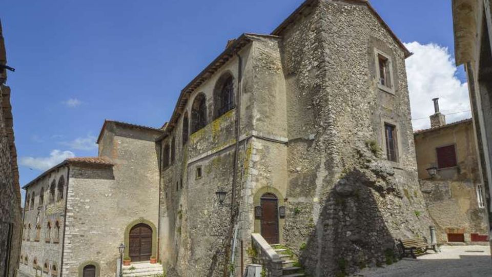 Antico edificio in pietra con facciata irregolare, grandi archi murati e finestre ad arco, affacciato su una piccola piazza lastricata con scalinate e muretti. Sullo sfondo si vedono altre case in pietra e un cielo azzurro con alcune nuvole.