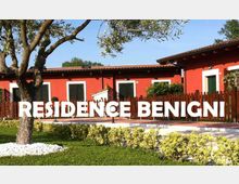 Edificio residenziale a un piano con facciata rossa, tetto in tegole e finestre con persiane verdi, circondato da una recinzione in legno. In primo piano ci sono un prato curato con aiuole e un albero, con la scritta “RESIDENCE BENIGNI” sovrapposta all’immagine.