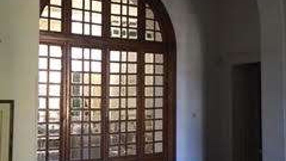Interno di un edificio con ampia porta finestra ad arco in legno e vetri a riquadri, affacciata su un cortile esterno. Pavimento a mosaico con motivi geometrici e soffitto alto con volta ad arco.