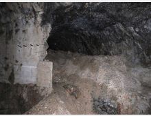 Interno di una galleria sotterranea in roccia grezza, con pareti irregolari e annerite e un piccolo passaggio ad arco che prosegue nel buio. Il pavimento è coperto da cumuli di terra e detriti, con un tratto di muratura o intonaco sul lato sinistro.