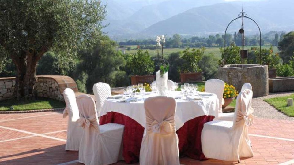 Tavolo rotondo apparecchiato all’aperto su una terrazza in mattoni, con sedie rivestite e tovaglia bianca e rossa. Sullo sfondo si vedono un giardino con alberi e vasi, un pozzo in pietra e una valle con colline e montagne.