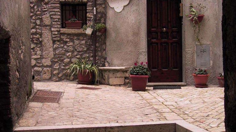 Cortile acciottolato visto da sotto un arco, con facciata di edificio in pietra e intonaco, porta in legno scuro e finestra con persiane. Sul muro c’è un’edicola con immagine e un cartello “Via Castello”, con vasi di fiori e piante disposti vicino all’ingresso.