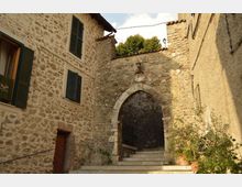 Vicolo in un borgo storico con edifici in pietra e persiane scure, che conduce a una scalinata. In fondo si vede un arco ogivale in muratura, simile a un’antica porta, incassato tra le pareti di pietra.