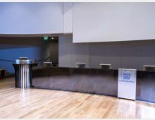 Sala conferenze moderna con ampio palco in legno e lungo banco relatori in metallo, con postazioni dotate di schermi. Ai lati sono presenti due podi, uno con il logo “Confindustria”, e una parete di fondo grigia sotto un soffitto alto.