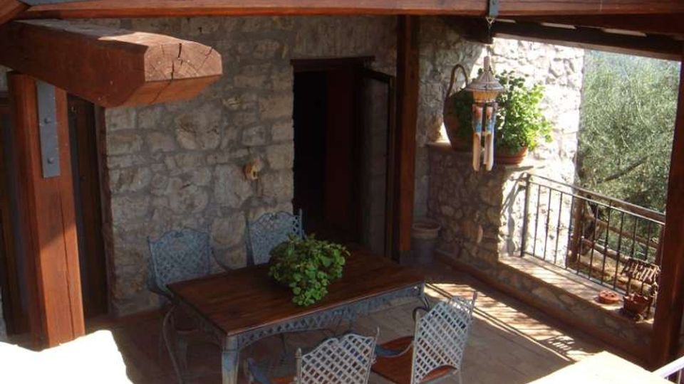 Terrazza coperta con travi in legno e pareti in pietra, arredata con tavolo e sedie in metallo. Sul lato destro ci sono ringhiera in ferro, vasi di piante e una lanterna sospesa, con vista sul verde circostante.