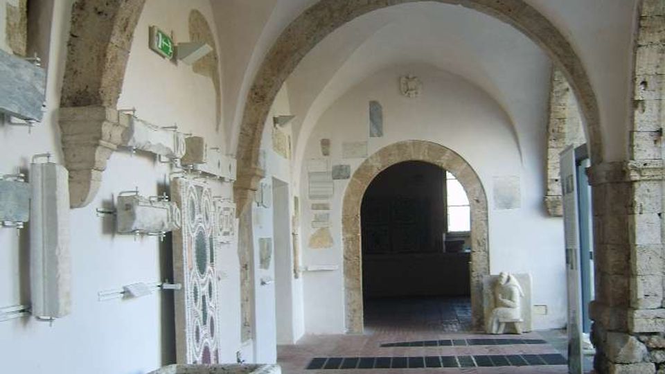 Corridoio interno con volte a botte e grandi archi in pietra su pareti intonacate bianche, pavimento in mattoni a spina di pesce e oggetti lapidei esposti lungo le pareti. In primo piano si vede una vasca in pietra, mentre sul fondo un arco conduce a un ambiente più scuro con una finestra.