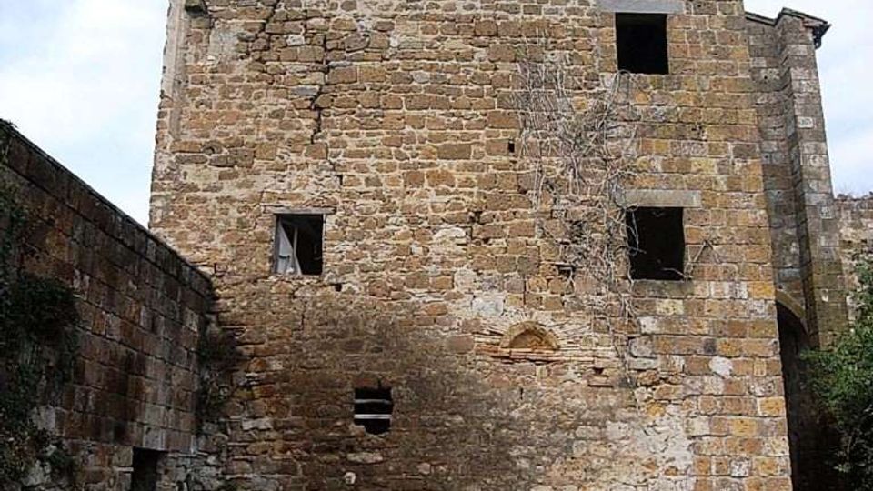 Edificio storico in pietra a tre livelli, con muratura irregolare, crepe visibili e finestre rettangolari prive di infissi. Davanti si vede un piccolo cortile in pietra delimitato da muri laterali, sotto un cielo parzialmente nuvoloso.
