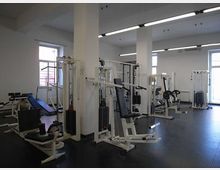 Interno di una palestra con pavimento scuro in gomma, grandi finestre e colonne bianche, illuminata da lunghe lampade a soffitto. Sono presenti varie macchine per allenamento con pesi e panche distribuite nella sala.