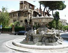 Fontana monumentale in una rotonda stradale, con grande vasca circolare e gruppo scultoreo in pietra attorno alla base. Sullo sfondo si vede una villa storica in stile eclettico con torre e logge, circondata da alberi alti e vegetazione urbana.
