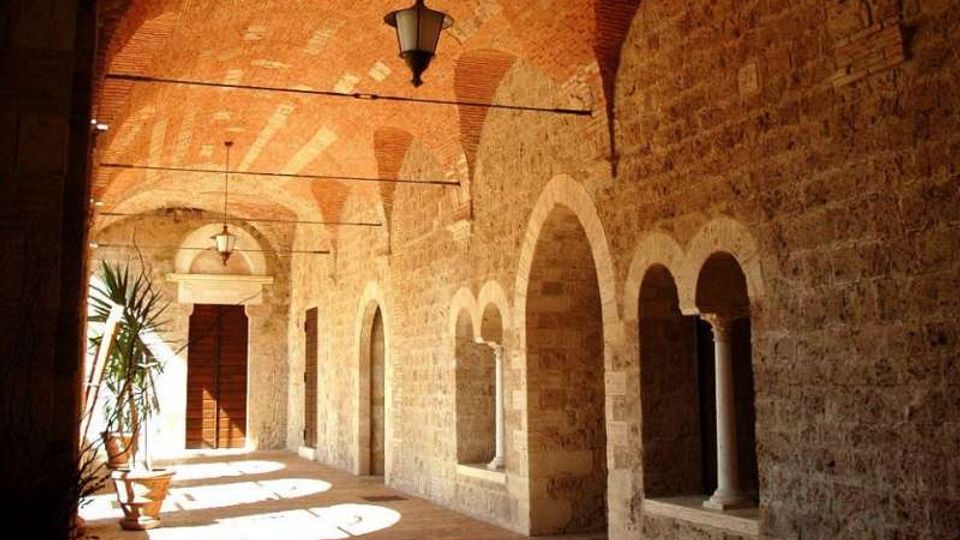Portico in stile storico con volta in mattoni e pareti in pietra, illuminato dalla luce laterale che crea ombre sul pavimento in cotto. Lungo il lato destro si susseguono arcate con colonne e nicchie, con lanterne sospese e alcune piante in vaso vicino all’ingresso sul fondo.