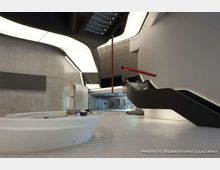 Interno di un edificio moderno con ampio atrio in cemento e superfici bianche, illuminato da luci integrate nel soffitto curvo. Due scale nere con corrimano rossi salgono ai piani superiori e al centro si trova un banco reception ovale bianco su pavimento lucido.